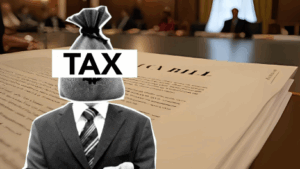 man in suit with a bag of money on his head, with the table tax. Background is what appears to be a written legislation bill sitting on a desk for approval.