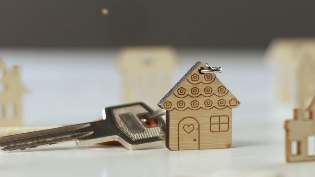 A close-up of a house-shaped wooden keychain attached to a silver key, resting on a white surface with blurred miniature house figures in the background.
