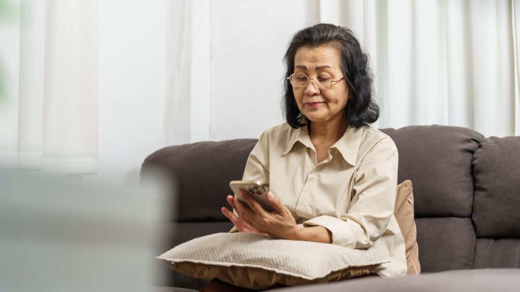 Elder Care - An older woman sits comfortably on a sofa, holding a smartphone and interacting with an AI eldercare companion app. She appears calm and engaged, using the technology in a relaxed home setting with soft natural light filtering through curtains in the background.