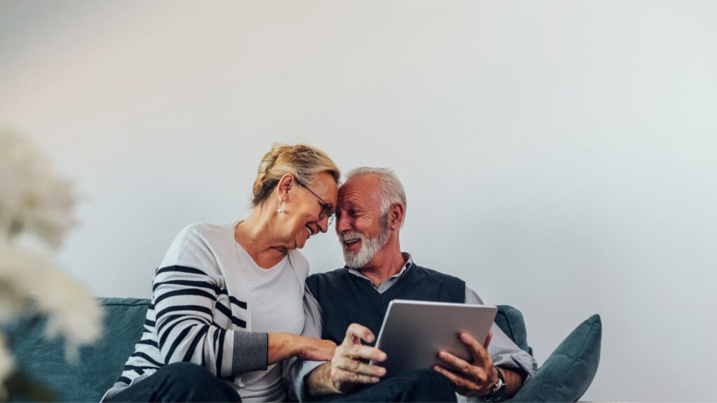Smiling older couple sitting on a couch, leaning their foreheads together while looking at a tablet, conveying comfort, connection, and confidence in their retirement planning.